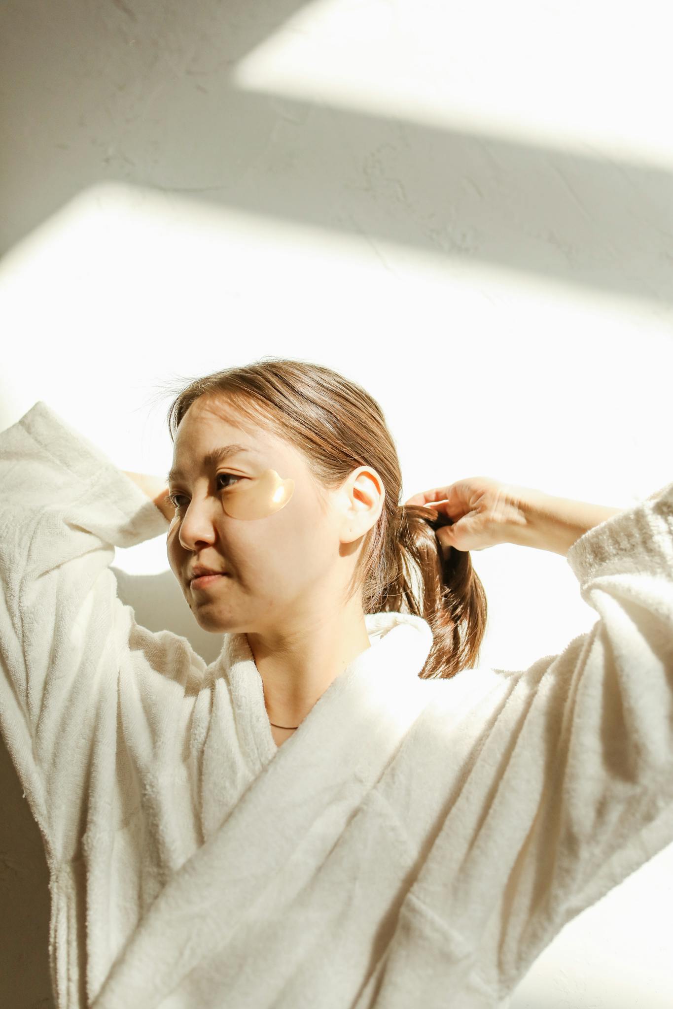 Asian woman in a bathrobe applying under eye masks as part of her skincare routine.