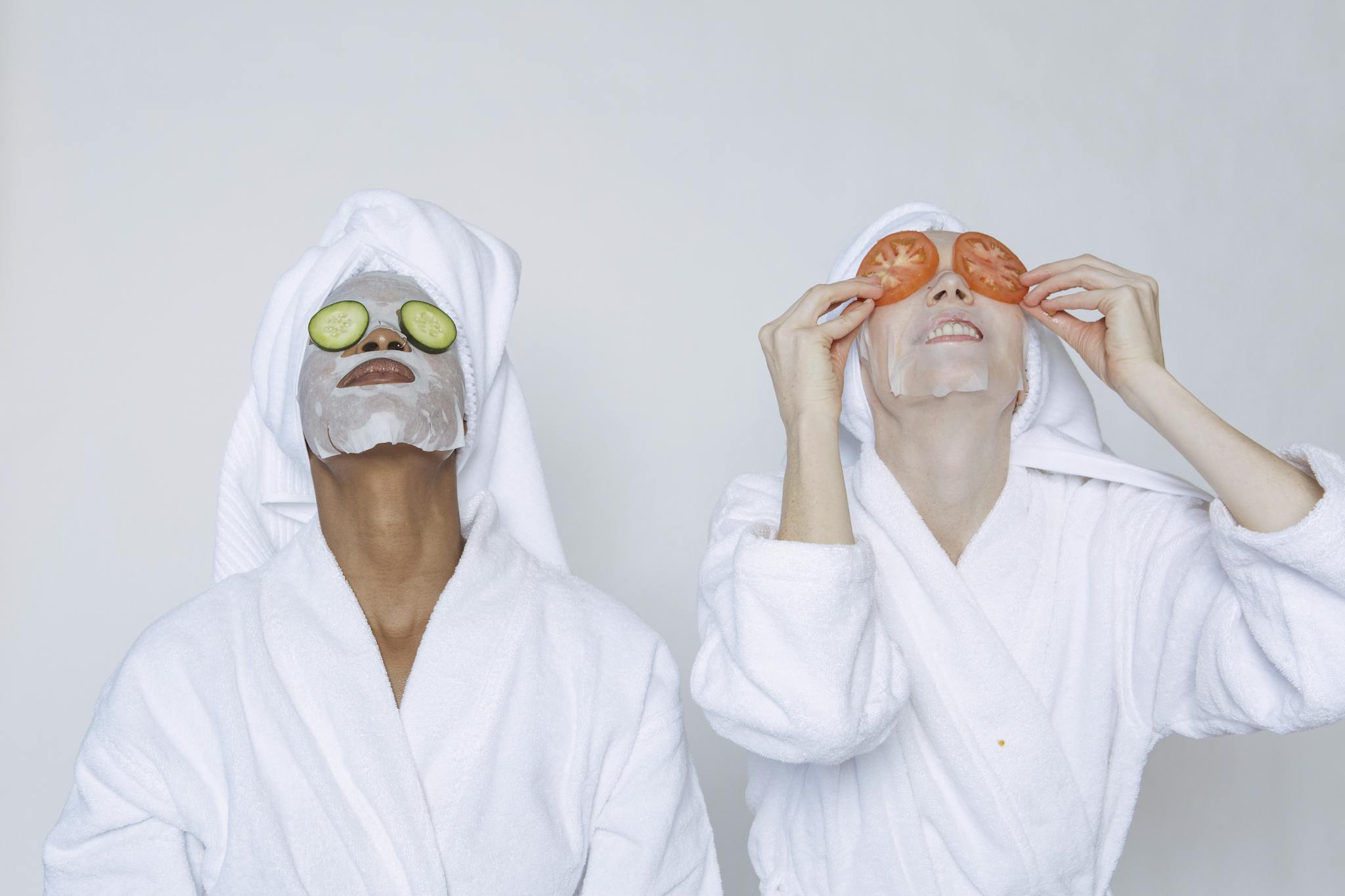 Young multiethnic females wearing bathrobes and towels moisturizing face with masks and slices of tomatoes and cucumbers against white background
