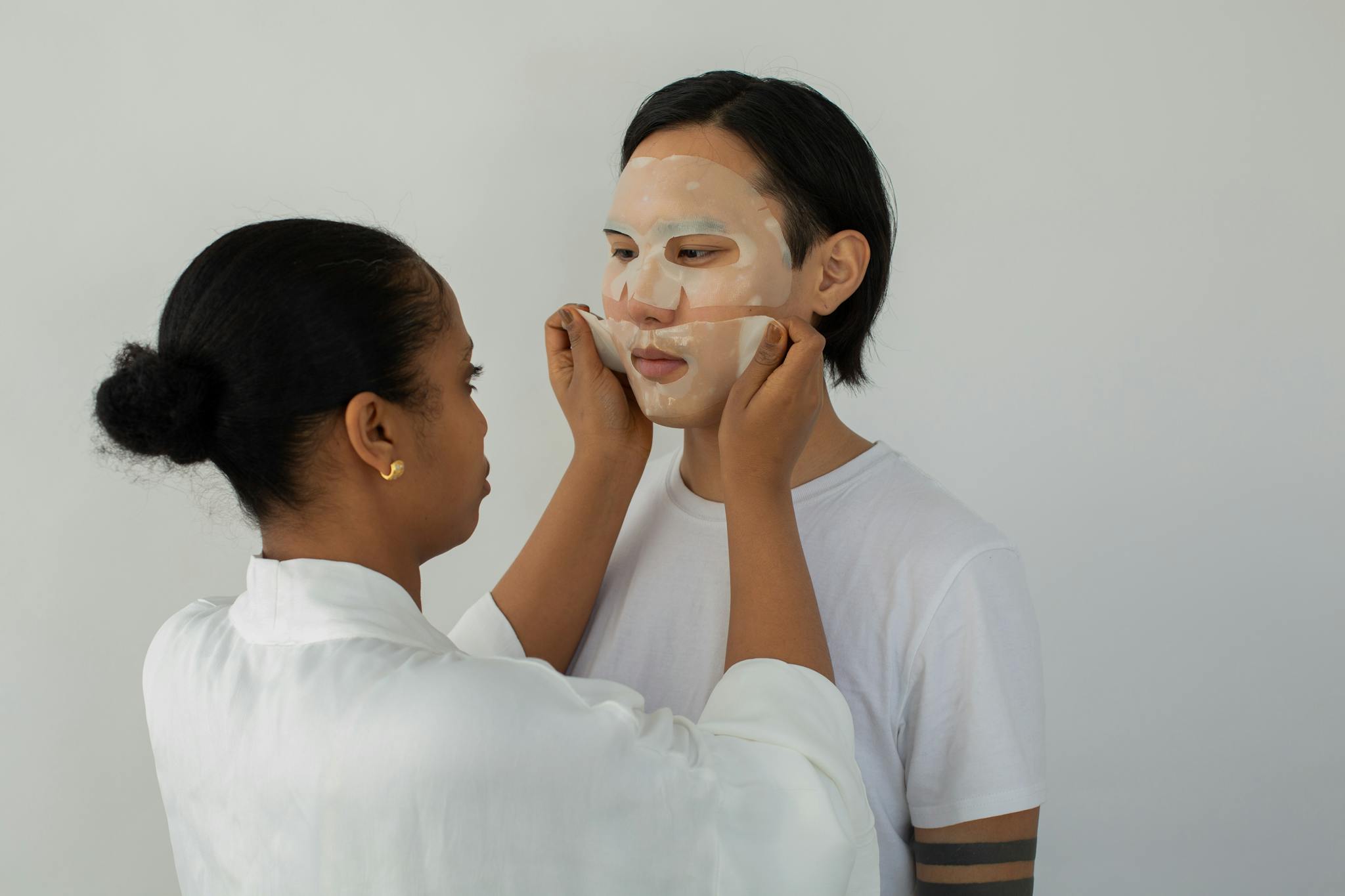 Black female cosmetician applying moisturizing mask on face of ethnic man while looking at each other on light background